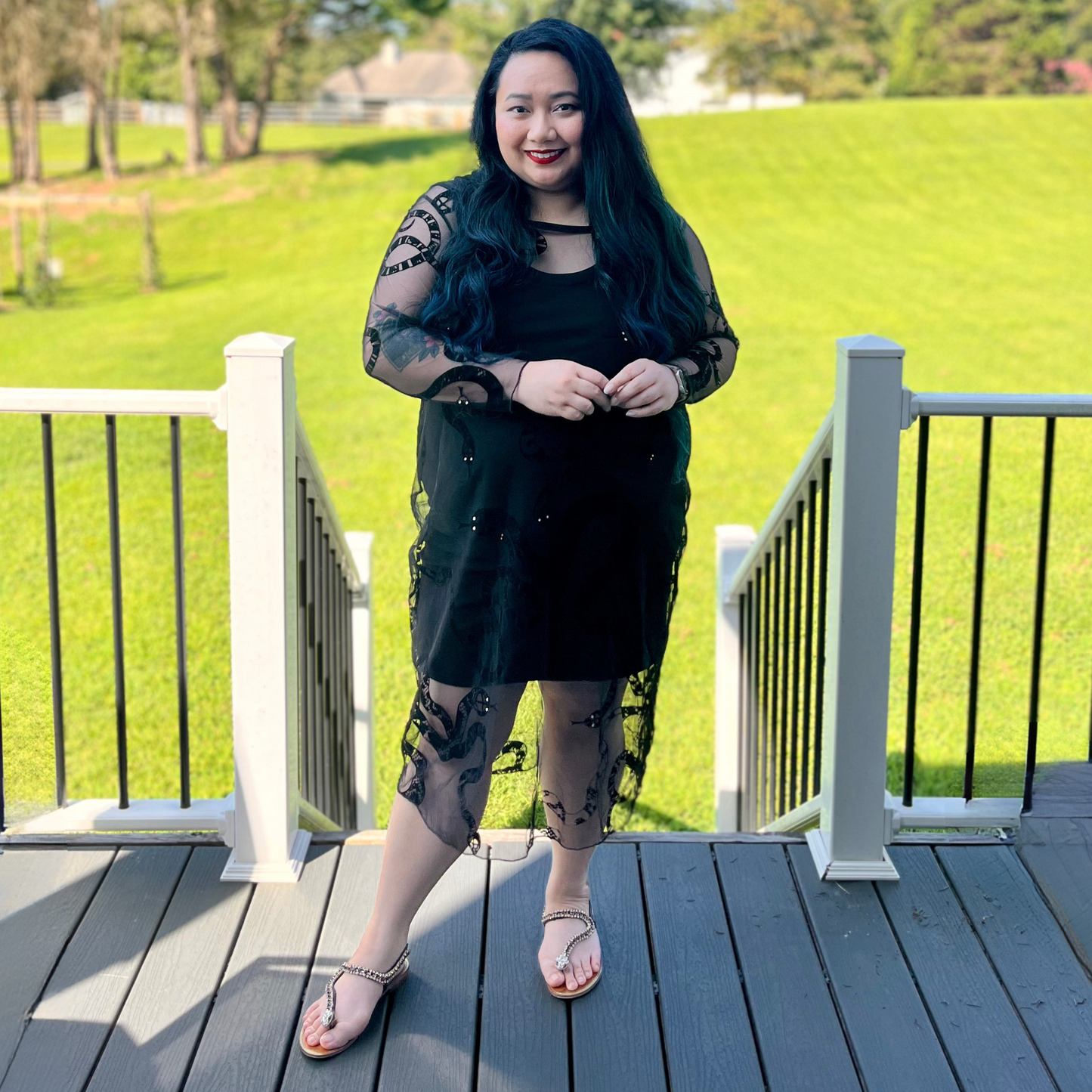 Woman in a black dress standing on a wooden deck with green grass and trees in the background