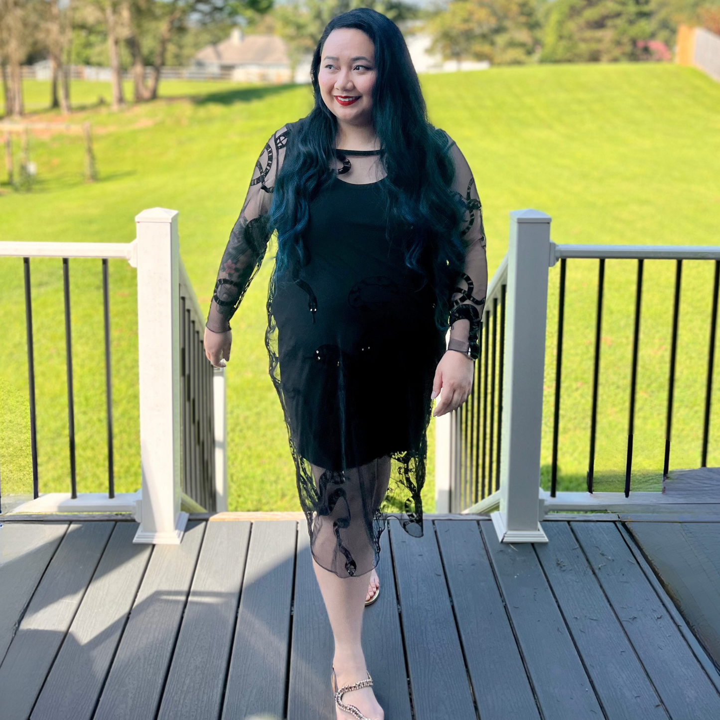 Woman in a black dress standing on a wooden deck with green grass and trees in the background