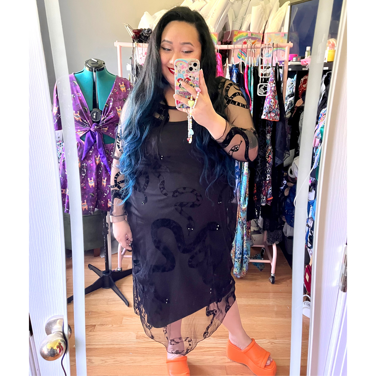 Woman taking a mirror selfie wearing a black dress with sheer overlay in a clothing store.