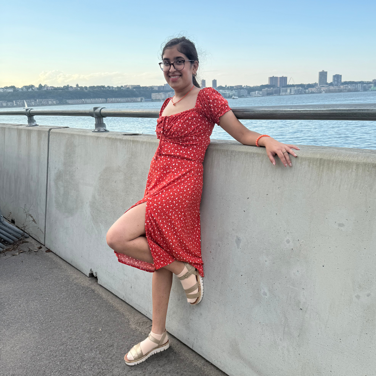 Woman in a red dress leaning against a concrete wall by a waterfront with a city skyline in the background.
