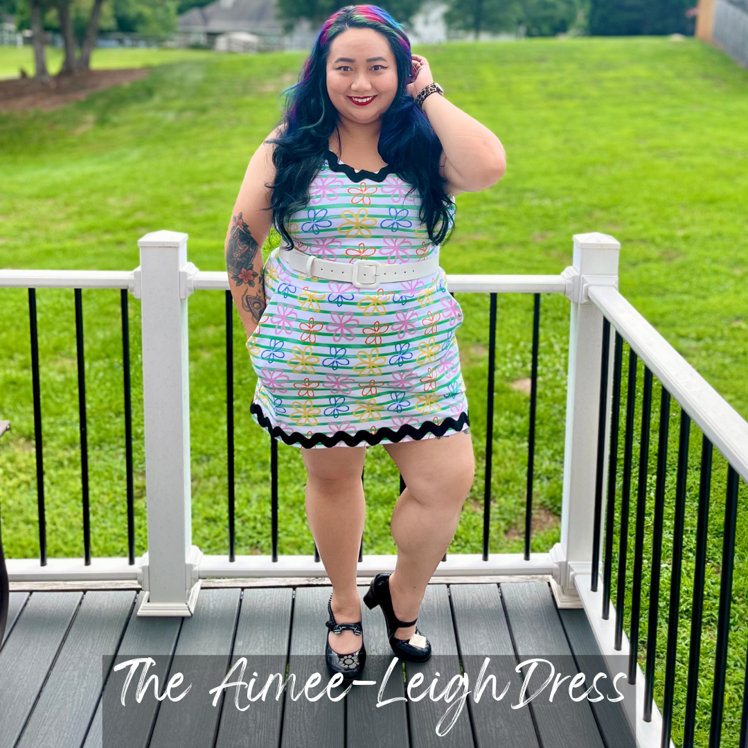 A woman stands on a white-railed deck outdoors wearing a colorful striped sleeveless dress with black trim, a white belt, and black heels. The background shows a green lawn.