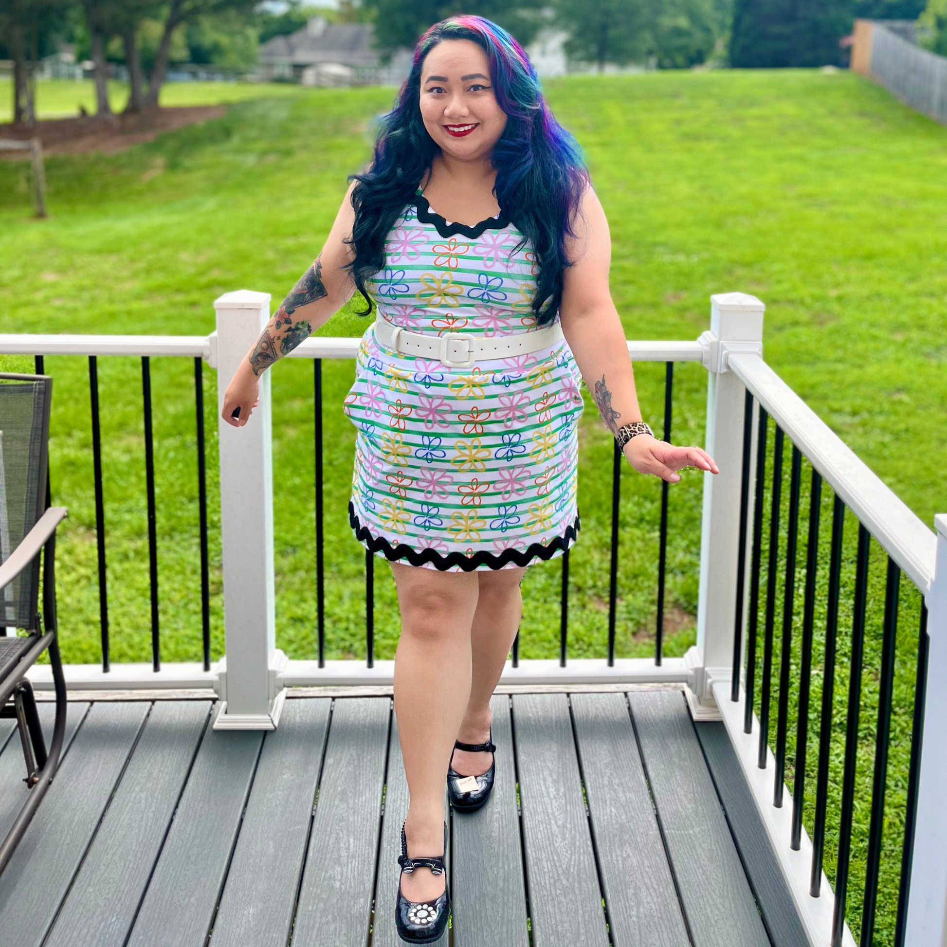 A woman with purple-dyed hair stands on a gray deck railing wearing a colorful striped mini dress with black trim and black Mary Jane-style shoes, with a green lawn in the background.