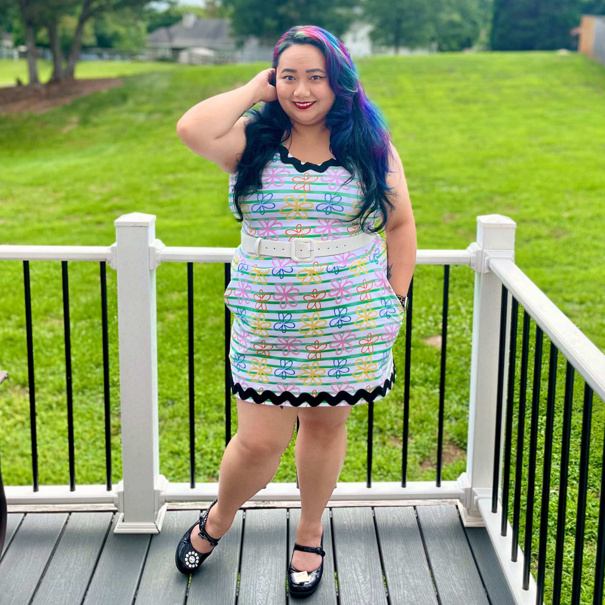 A woman stands on a wooden deck wearing a colorful, patterned sleeveless mini dress with a white belt. The background shows a green grassy lawn and trees under daylight.