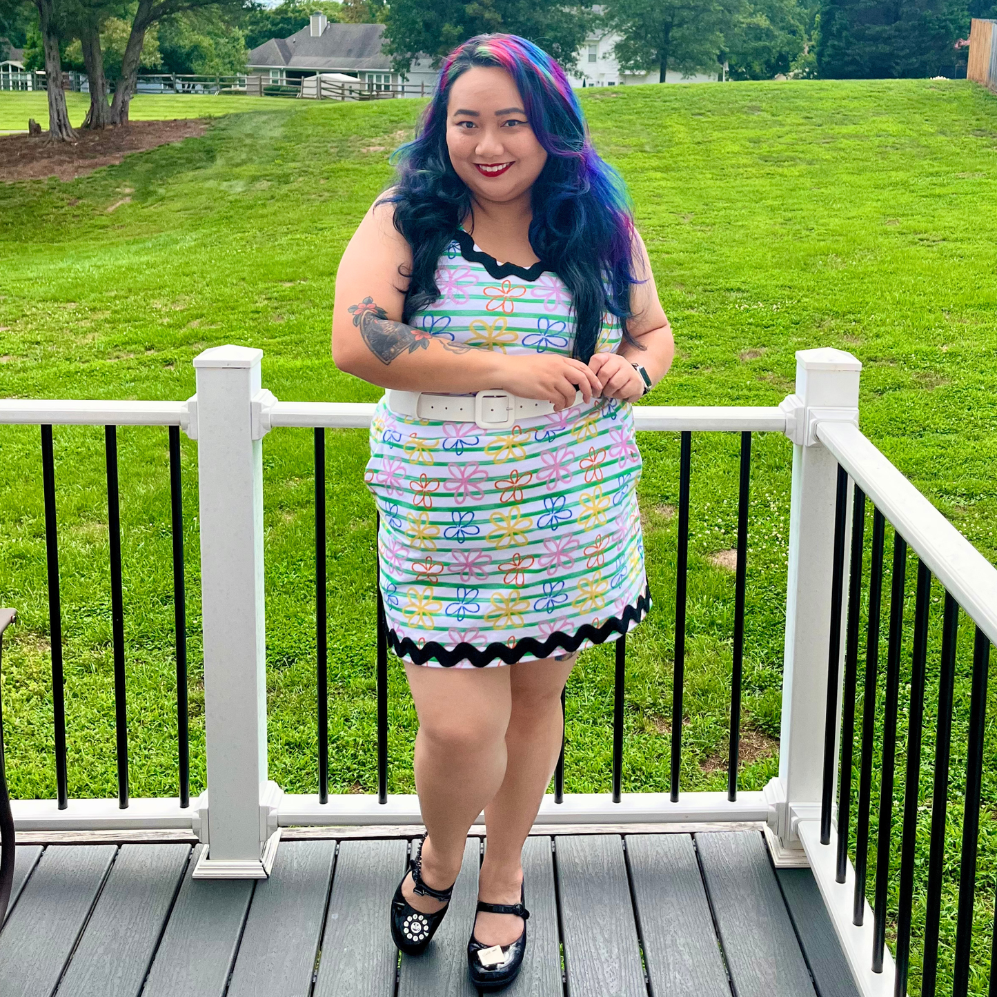 A woman stands on a wooden deck wearing a colorful patterned sleeveless mini dress with black trim and a white belt, photographed outdoors with a grassy lawn and trees in the background.