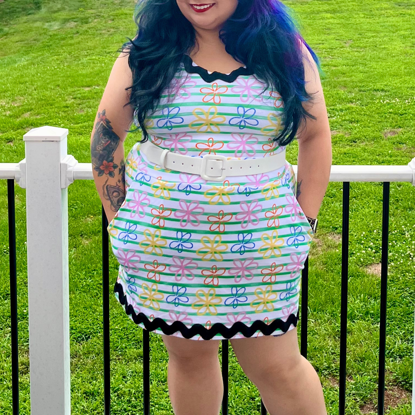 A woman wearing a sleeveless white mini dress with a colorful floral pattern, black scalloped trim, and a white belt, standing outdoors in front of a white fence and green grass.