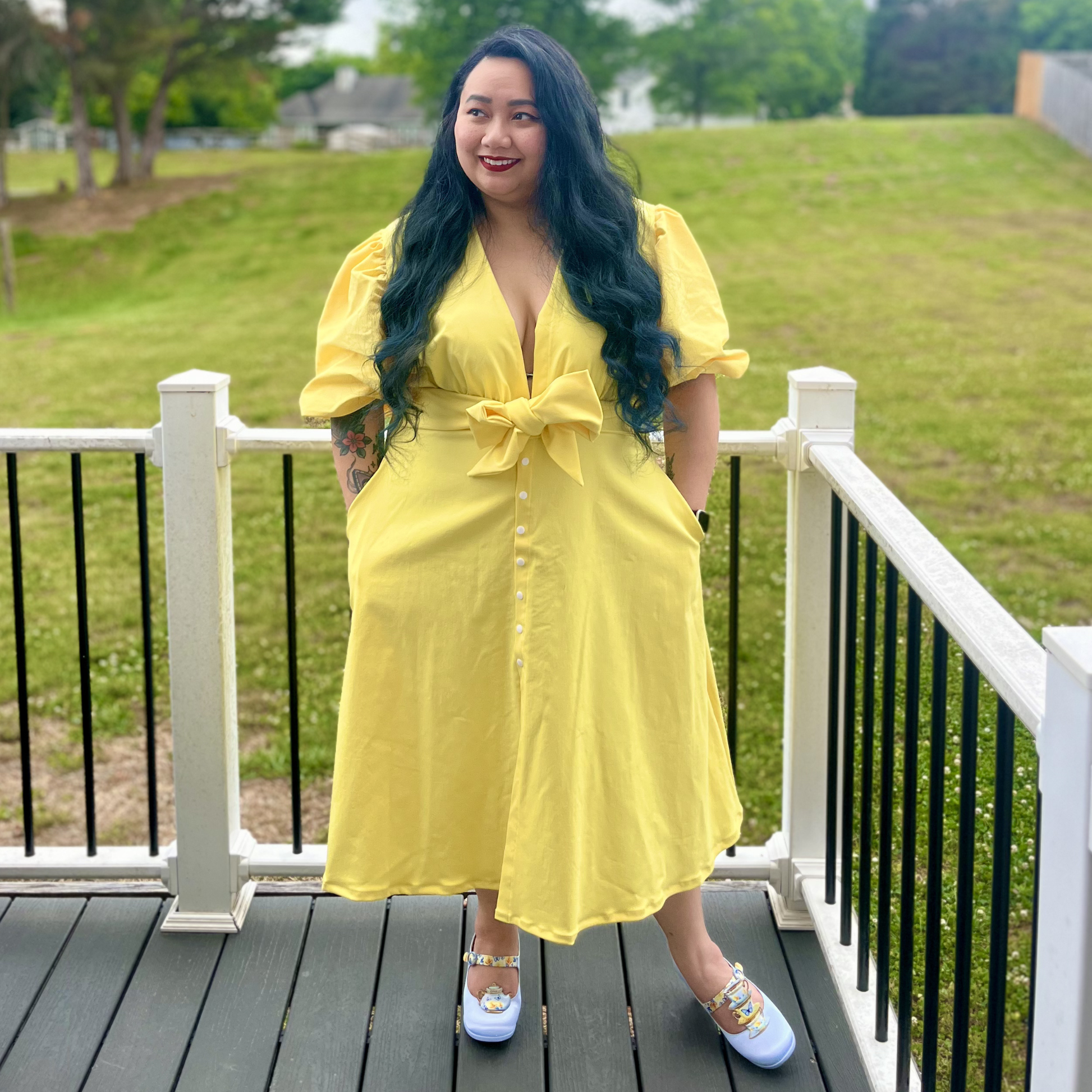 Woman in a yellow dress standing on a deck with green grass and trees in the background