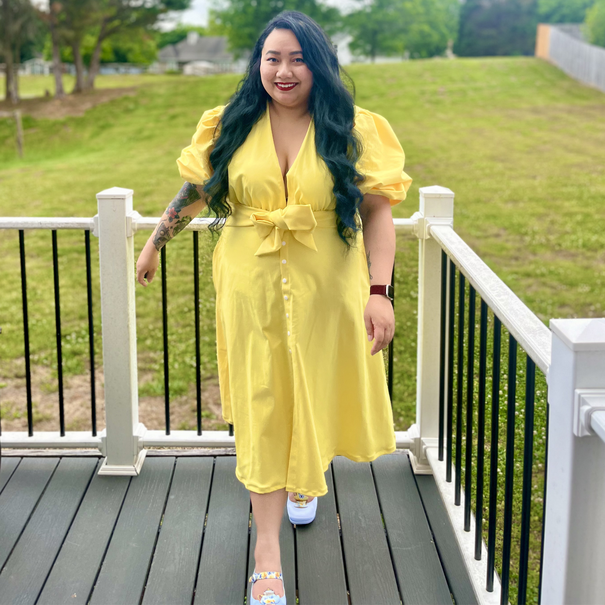 Woman in a yellow dress standing on a wooden deck with grass and trees in the background