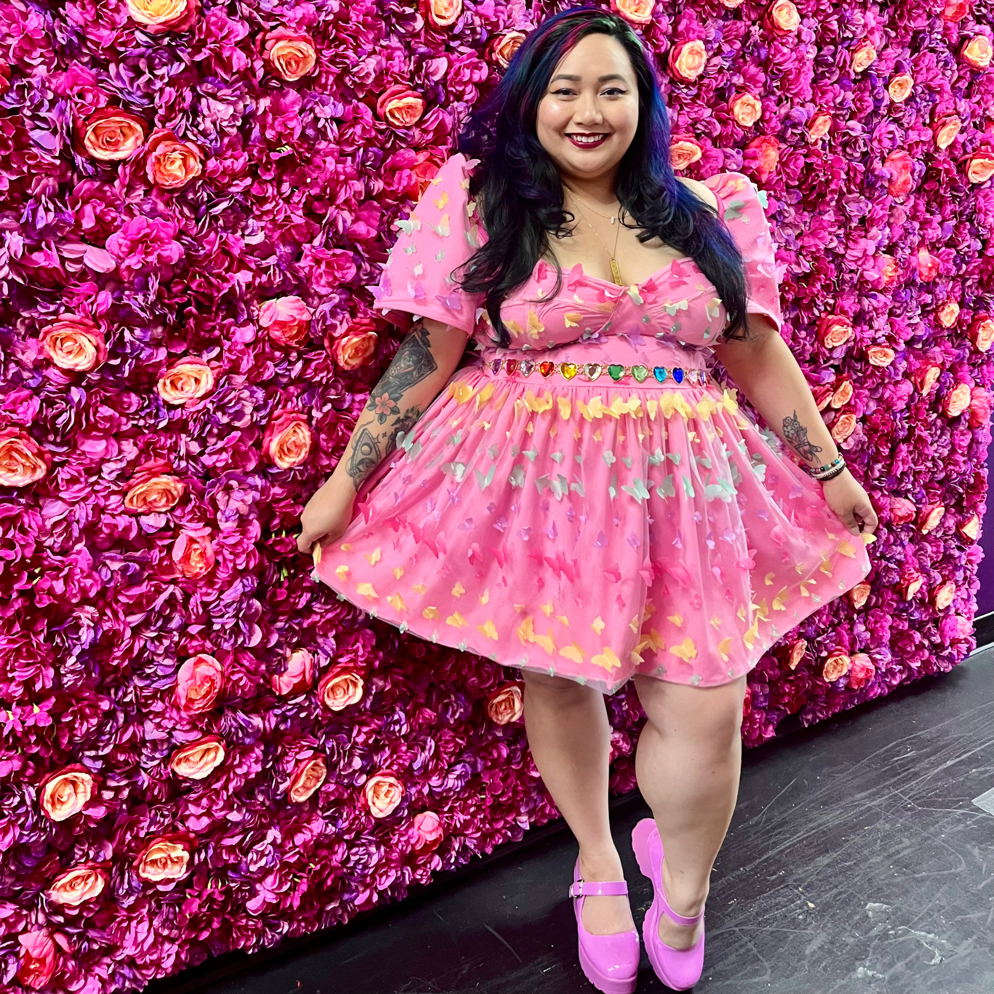 A woman posing in a pink short puff-sleeve dress with rainbow butterfly details standing in front of a vibrant pink flower wall.
