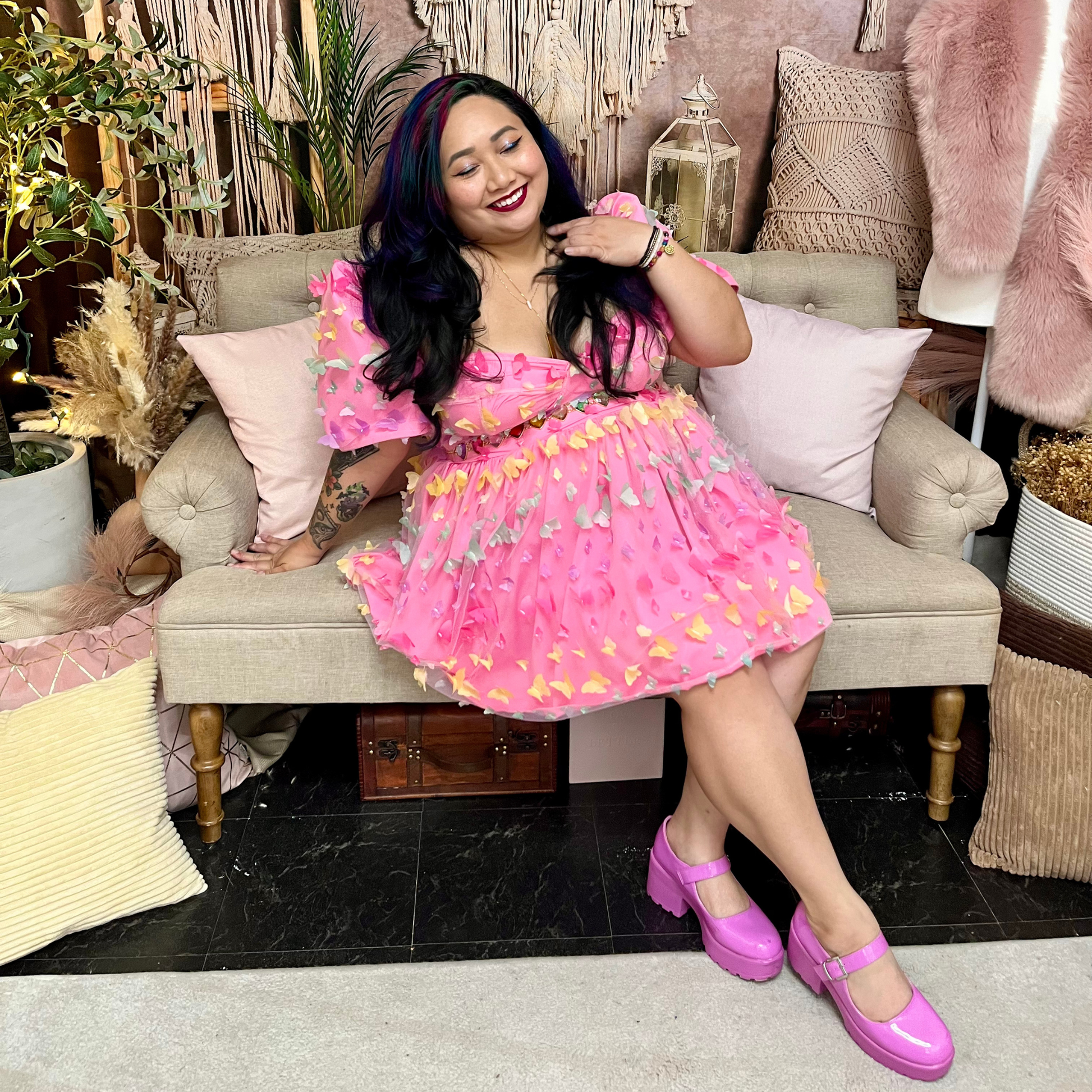 A woman wearing a pink mini dress with a colorful patterned skirt and bright pink Mary Jane shoes, sitting on a beige sofa in a decorated room.