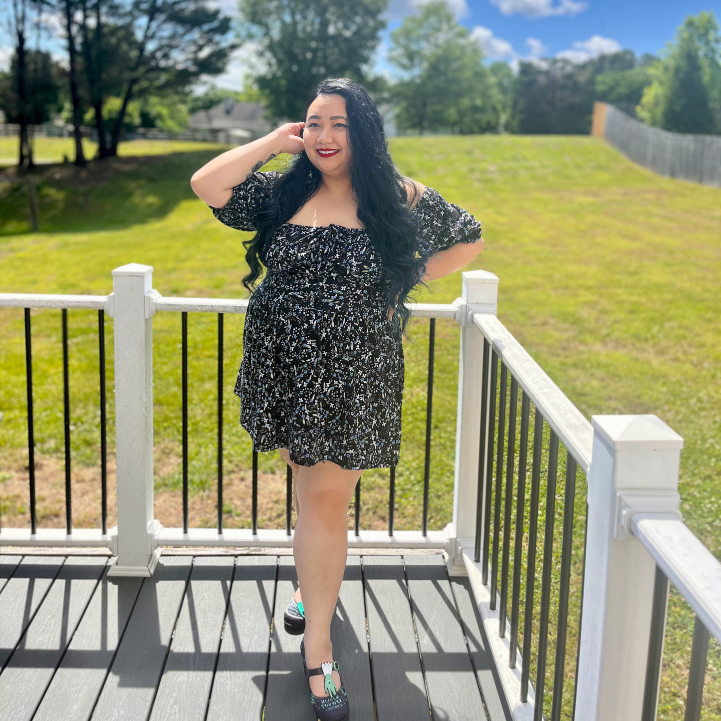Woman wearing a floral dress standing on a deck in front of grass and trees
