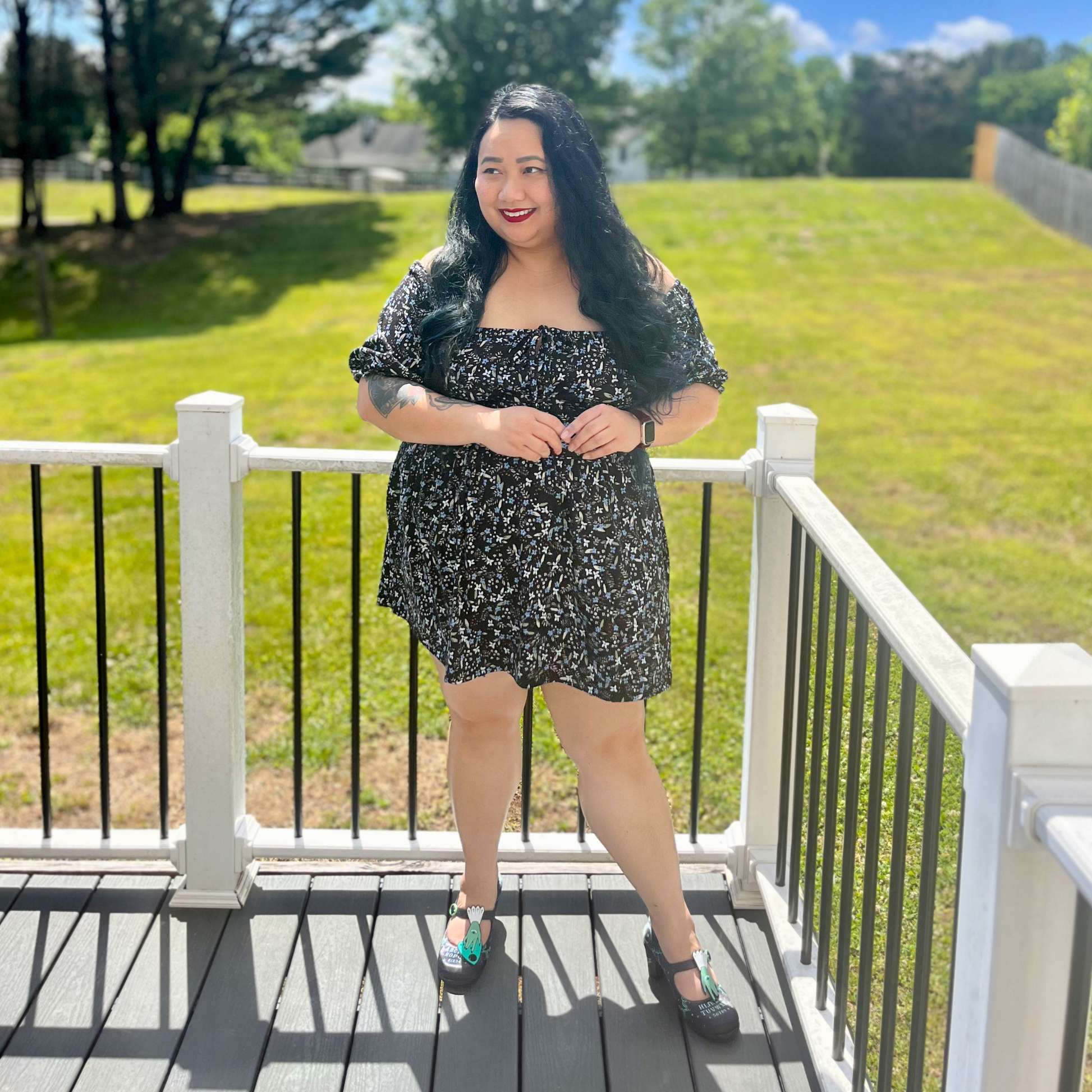 Woman wearing a floral dress standing on a deck in front of grass and trees