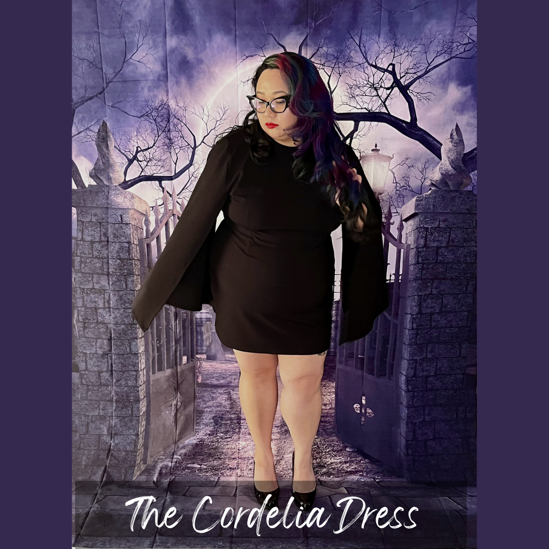 A woman wearing a black long-sleeve mini dress with dramatic draped sleeves stands in front of a gothic stone gate backdrop. The text 'The Cordelia Dress' appears at the bottom.