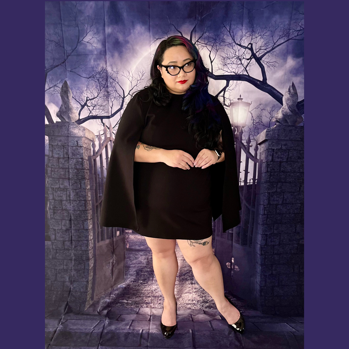 A woman modeling a black mini dress with a matching black cape, standing in front of a gothic-themed backdrop depicting a stone gate, bare trees, and a cloudy purple sky.
