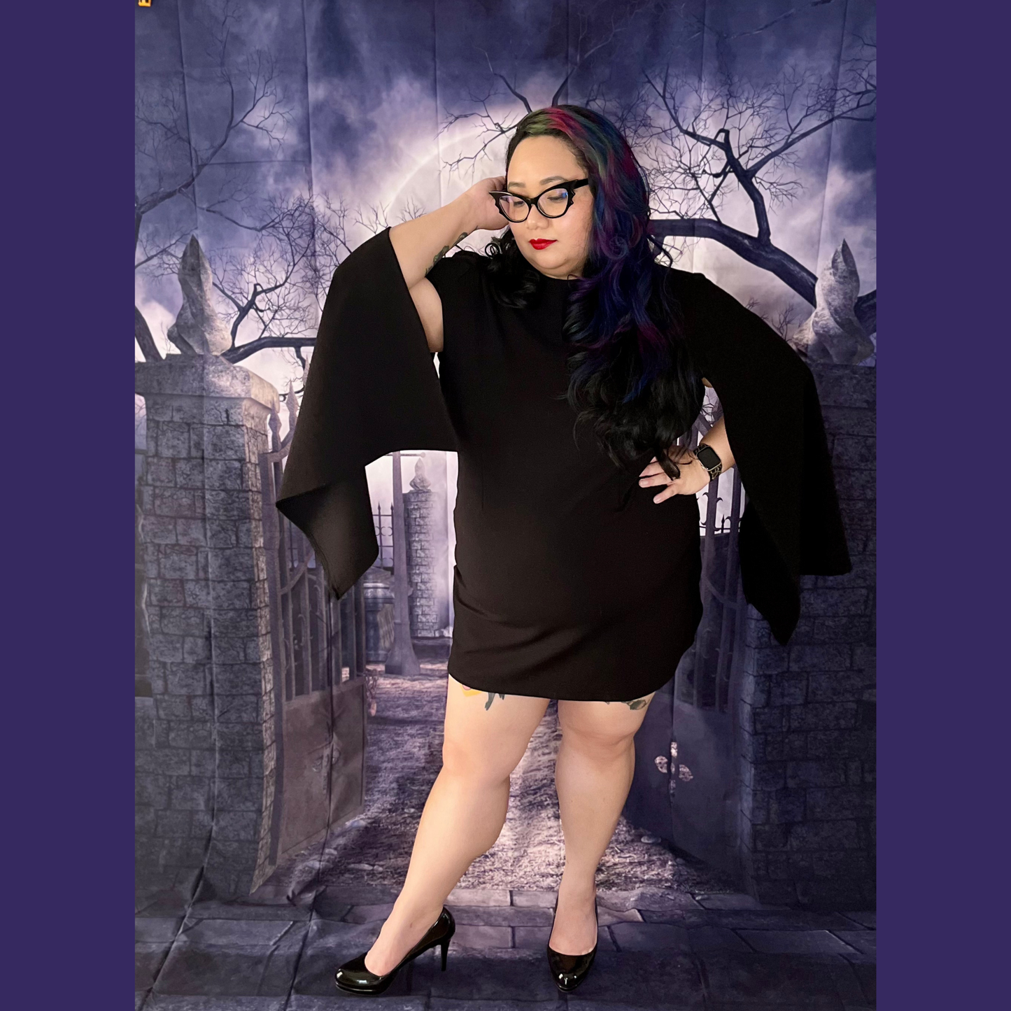 A woman modeling a black mini dress with dramatic wide cape sleeves, posed in front of a gothic stone alley backdrop with a full moon and bare trees.