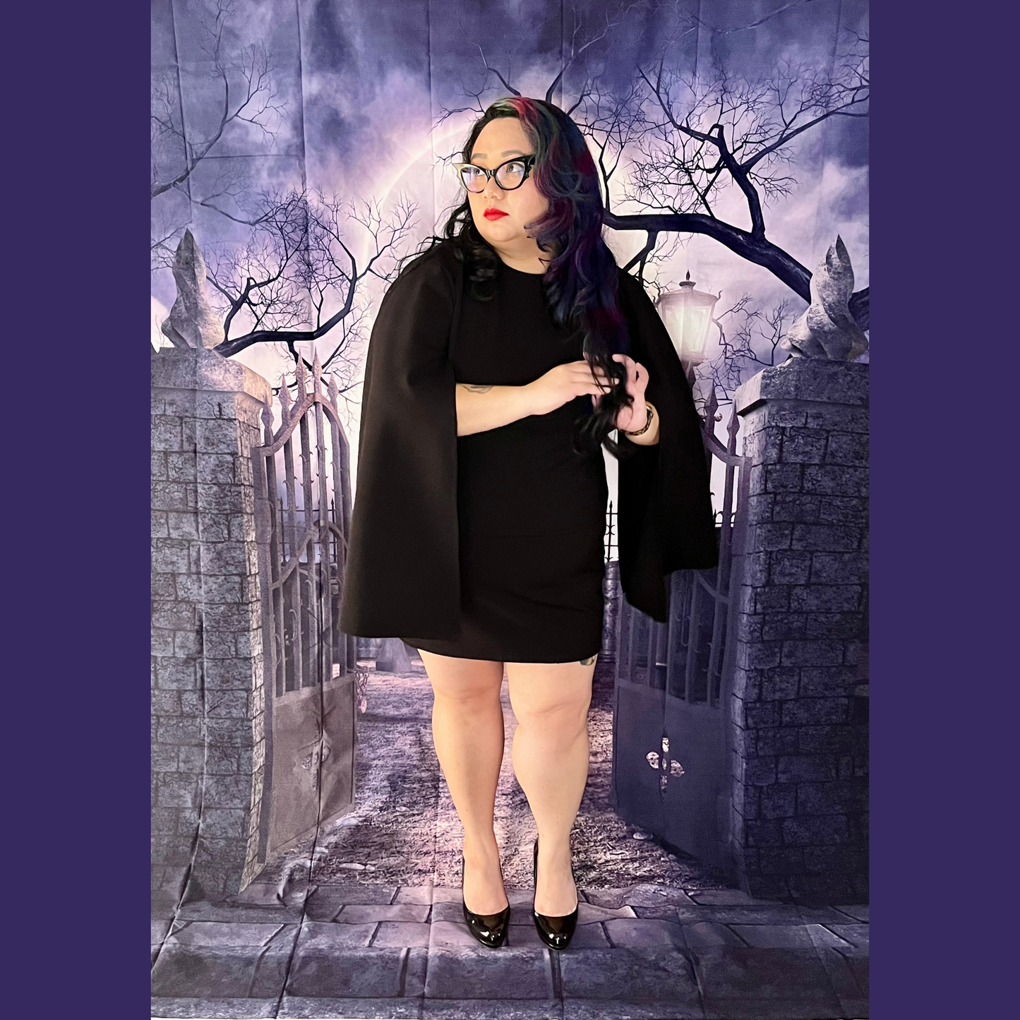 A woman wearing a black long-sleeve mini dress with dramatic draped sleeves stands in front of a gothic stone gate backdrop. 