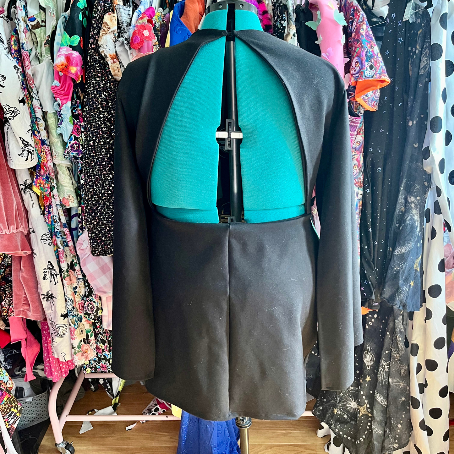 A black tailored jacket displayed on a dress form from the back, set against a background of colorful clothing racks.