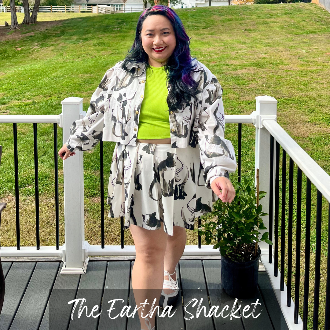 A woman stands on a wooden deck wearing a matching two-piece outfit from The Earth Shacket, featuring a black and white cat print jacket and skirt over a bright lime green crop top. The background shows a grassy lawn and white railings.