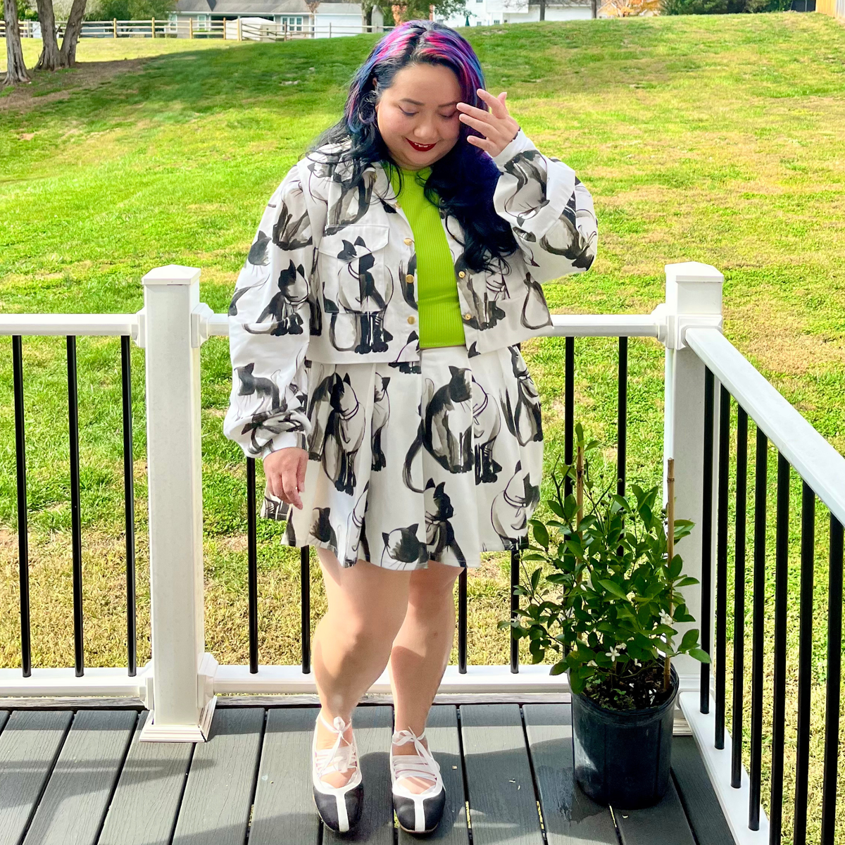 A person stands on a wooden deck outdoors wearing a white cropped jacket and matching mini skirt with a black cat print, and a bright lime green top. A potted plant is beside them, and a grassy lawn is in the background.