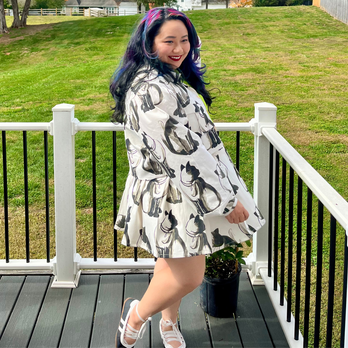 A woman with purple and blue hair stands on a wooden deck wearing a white long-sleeve jacket and skirt with a black cat pattern, posing outdoors with a grassy lawn in the background.