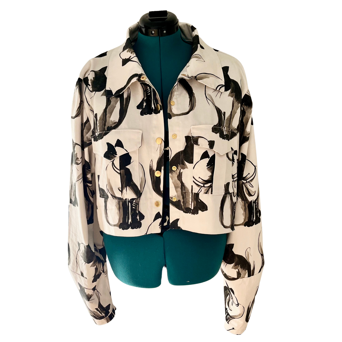 A cropped button-up shirt jacket featuring an all-over abstract black and gray cat print on a cream background, displayed on a dark teal mannequin torso against a white background.