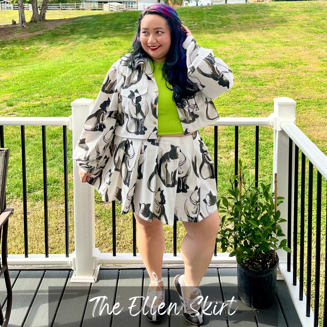 A woman stands on a wooden deck wearing a white matching jacket and skirt set with a black cat print, layered over a bright lime green top. The background shows a grassy lawn and white railings.