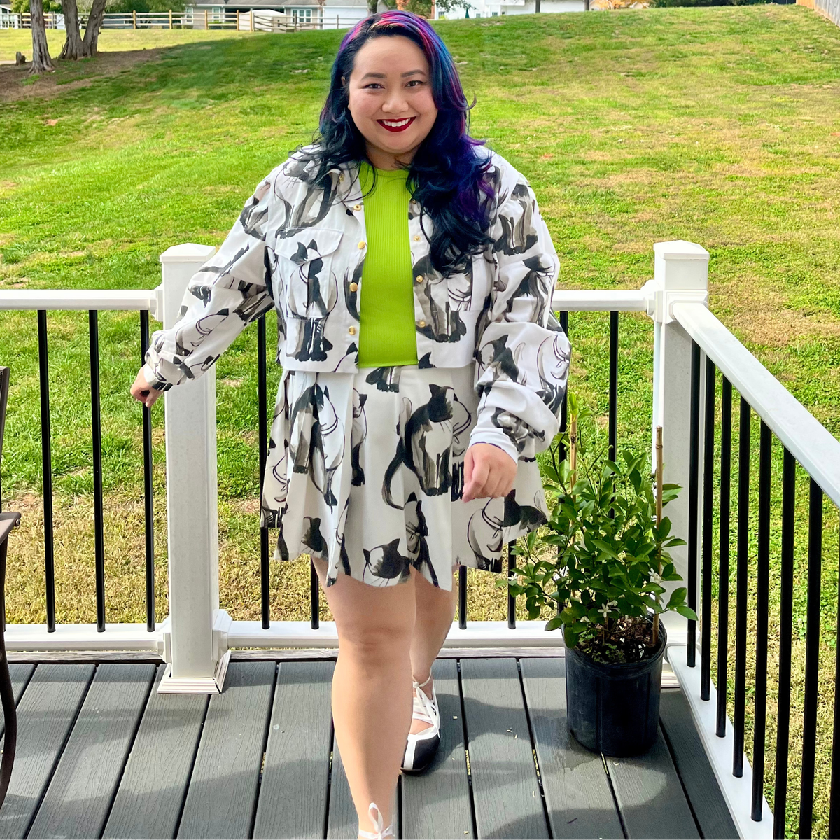 A woman stands on a wooden deck wearing a matching white, black, and gray abstract-patterned jacket and skirt set over a bright lime green top. The background shows a grassy lawn and white railings.