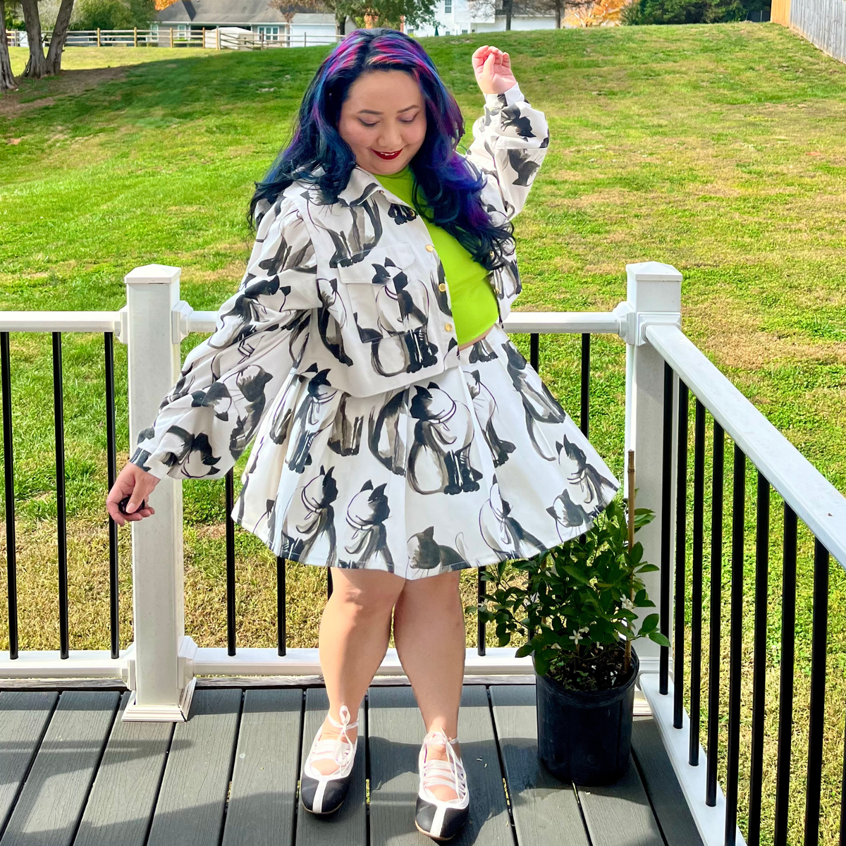 A person stands on a wooden deck wearing a white long-sleeve jacket and matching skirt set with a black cat print, layered over a bright neon green top. The background shows a grassy lawn and outdoor railing.