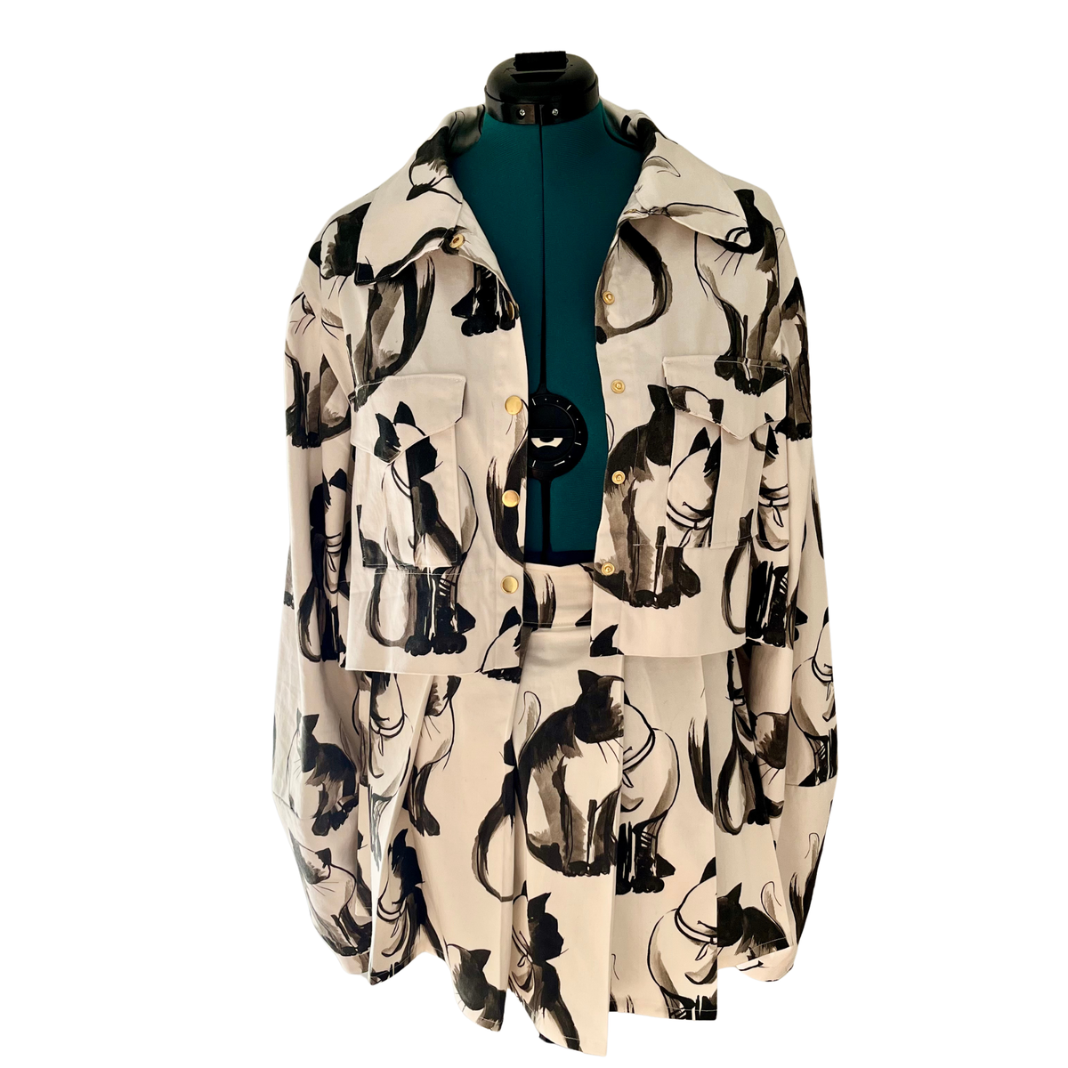 A cropped button-up shirt jacket  and skirt with a black and beige cat print, displayed on a dark green mannequin torso against a white background.