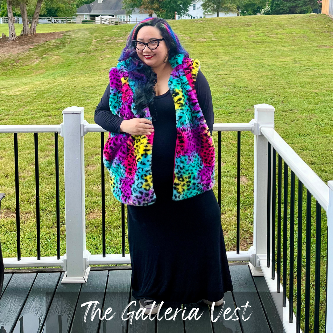 A woman wearing a vibrant, multicolor leopard-print vest stands on a wooden deck with a white railing, with a green grassy hill in the background. The text 'The Galleria Vest' is written at the bottom.