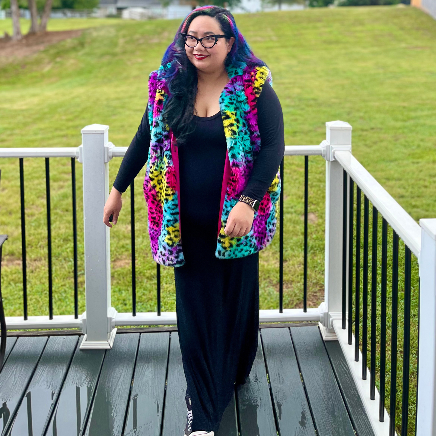 A woman stands on a wooden deck wearing a bright multicolor leopard-print faux fur vest over a black outfit, with a grassy lawn in the background.
