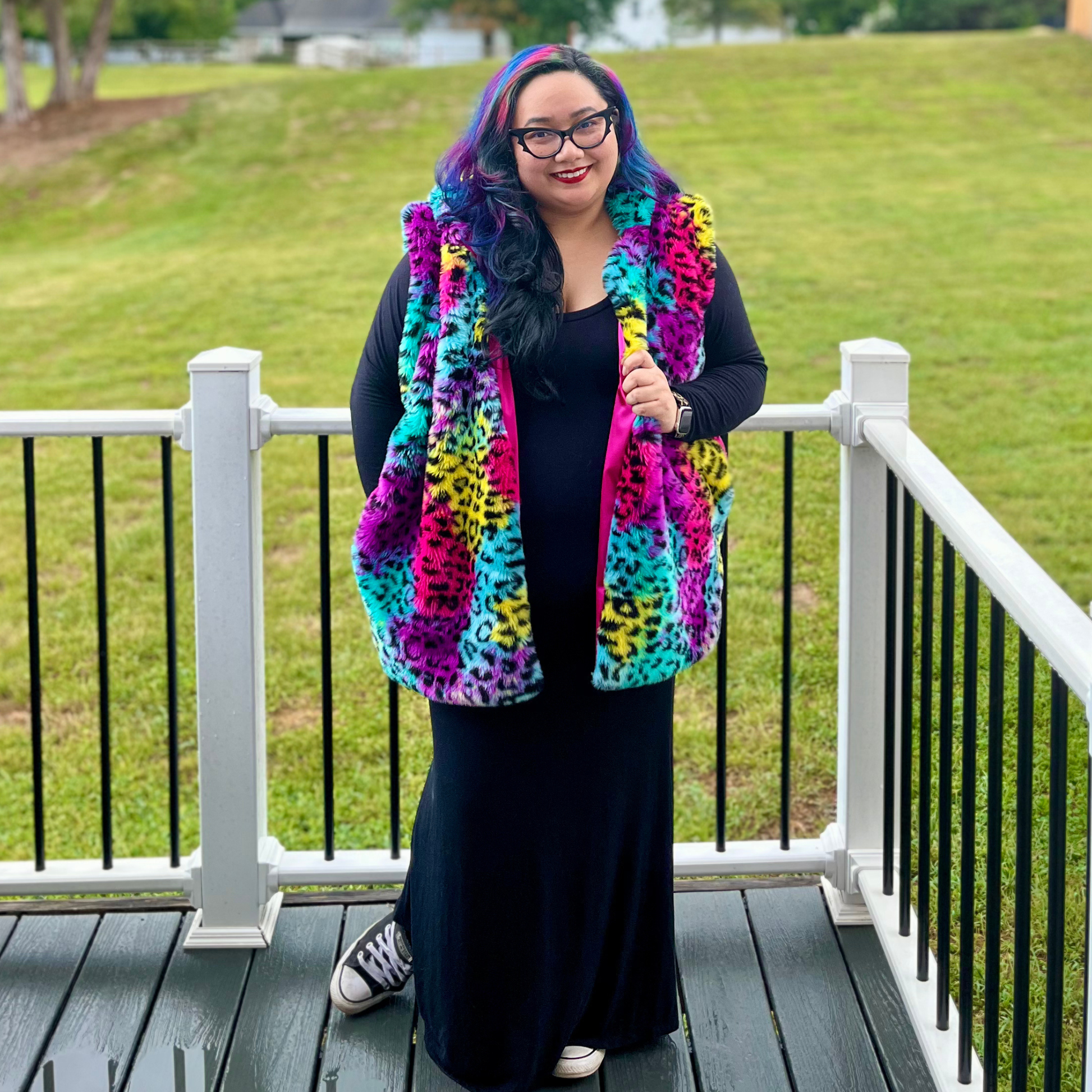 A woman stands on a wooden deck wearing a bright multicolor fuzzy vest with a leopard print pattern in pink, yellow, blue, and purple over a black outfit. The background shows a grassy lawn and white railings.