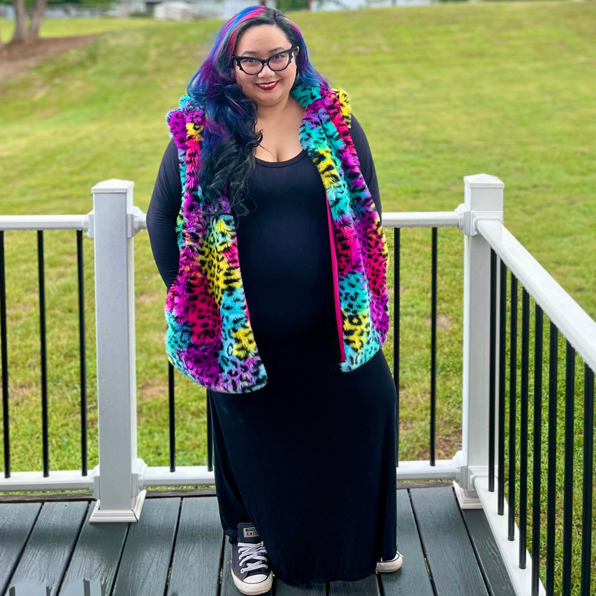 A woman with colorful blue, purple, and pink hair stands on a wooden deck wearing a vibrant, multicolored fuzzy vest with a pink leopard print over a black dress. The background shows a green grassy lawn behind a white railing.
