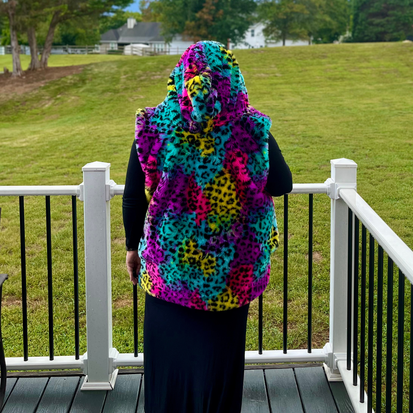 A person standing on a white-railled deck overlooking a grassy lawn, wearing a hooded fuzzy vest with a bright multicolor leopard print pattern in turquoise, pink, purple, yellow, and black.