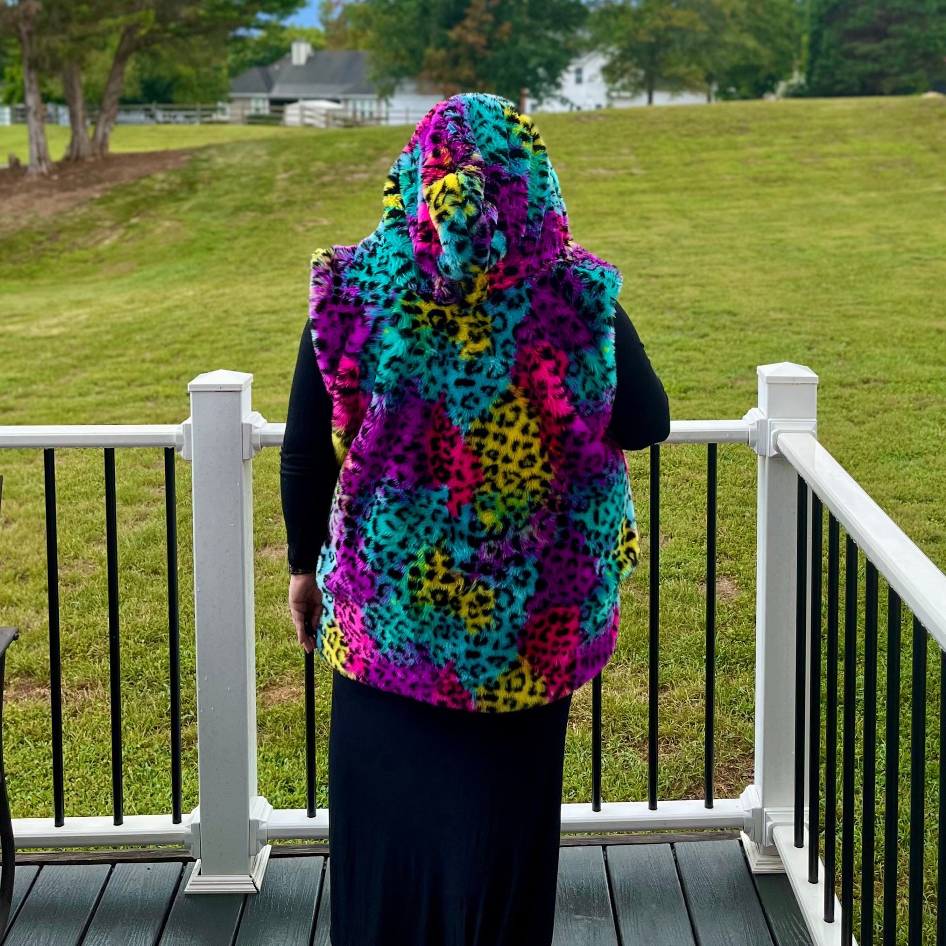 A person standing on a white-railled deck overlooking a grassy lawn, wearing a hooded fuzzy vest with a bright multicolor leopard print pattern in turquoise, pink, purple, yellow, and black.