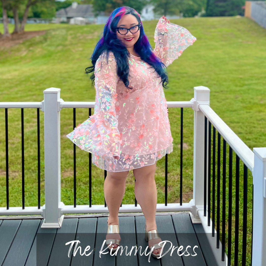 A woman with blue and purple hair stands on a white-railed deck wearing a sheer iridescent pink dress with floral print and flared sleeves. The text 'The Kimmy Dress' is overlaid at the bottom.