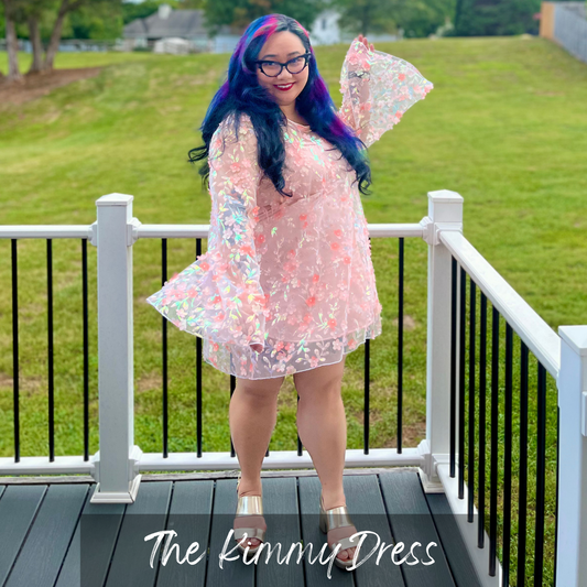 A woman with blue and purple hair stands on a white-railed deck wearing a sheer iridescent pink dress with floral print and flared sleeves. The text 'The Kimmy Dress' is overlaid at the bottom.