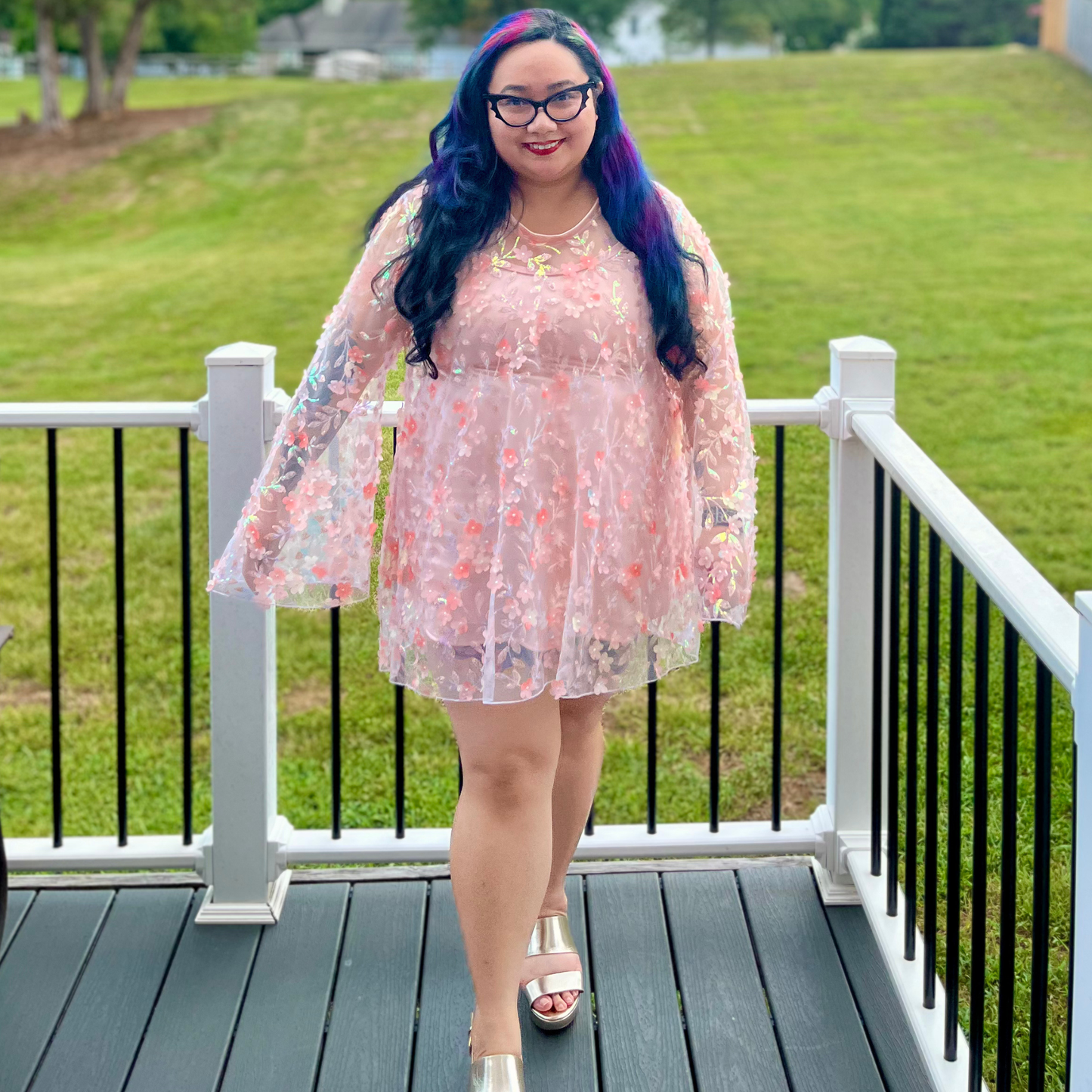 A woman with purple and blue hair stands on a wooden deck wearing a sheer light pink floral tunic dress with long bell sleeves and silver sandals, with a grassy lawn in the background.