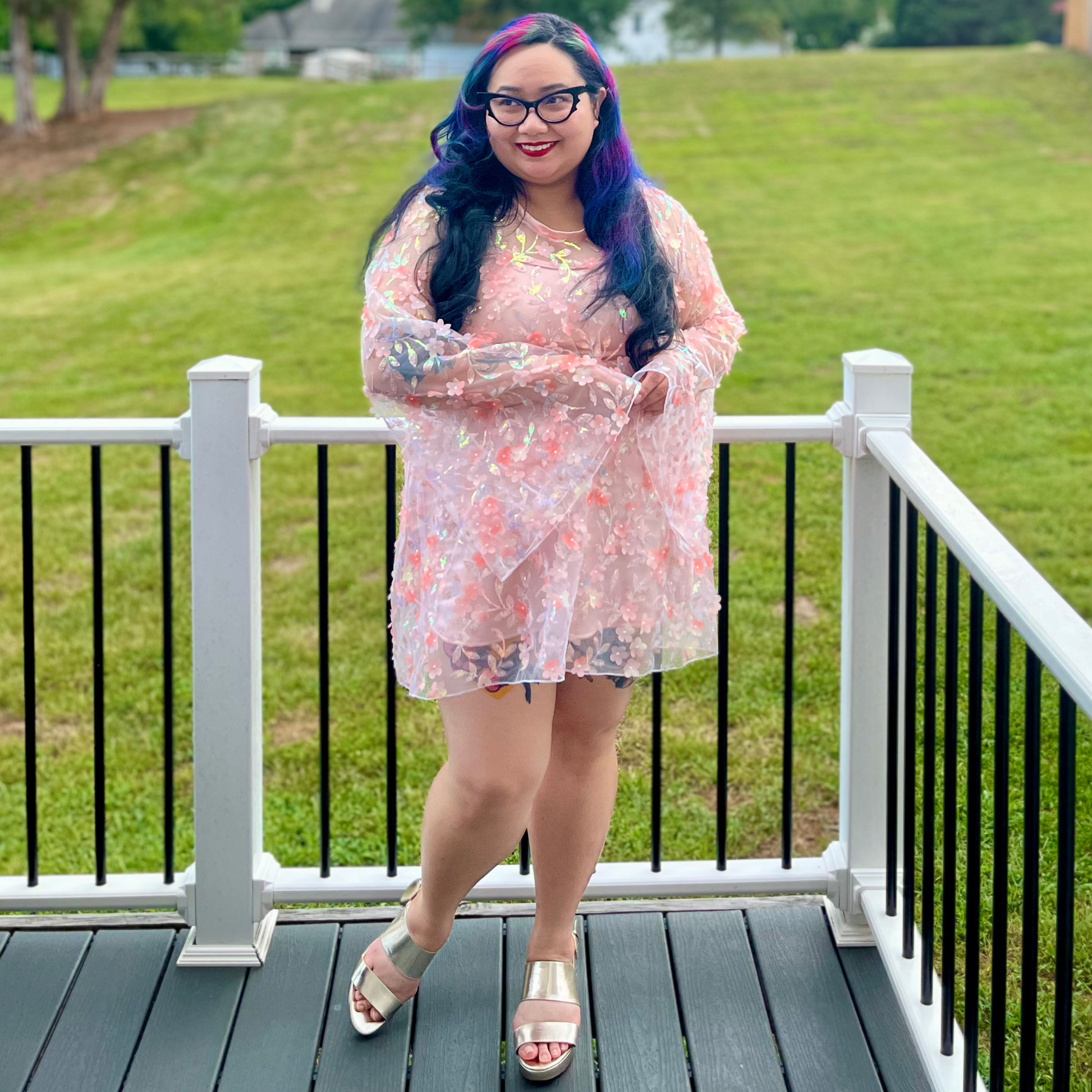 A woman with long, dark blue and purple hair stands on a wooden deck wearing a sheer, light pink floral long-sleeve dress with a matching headband and silver sandals, with a green grassy field in the background.