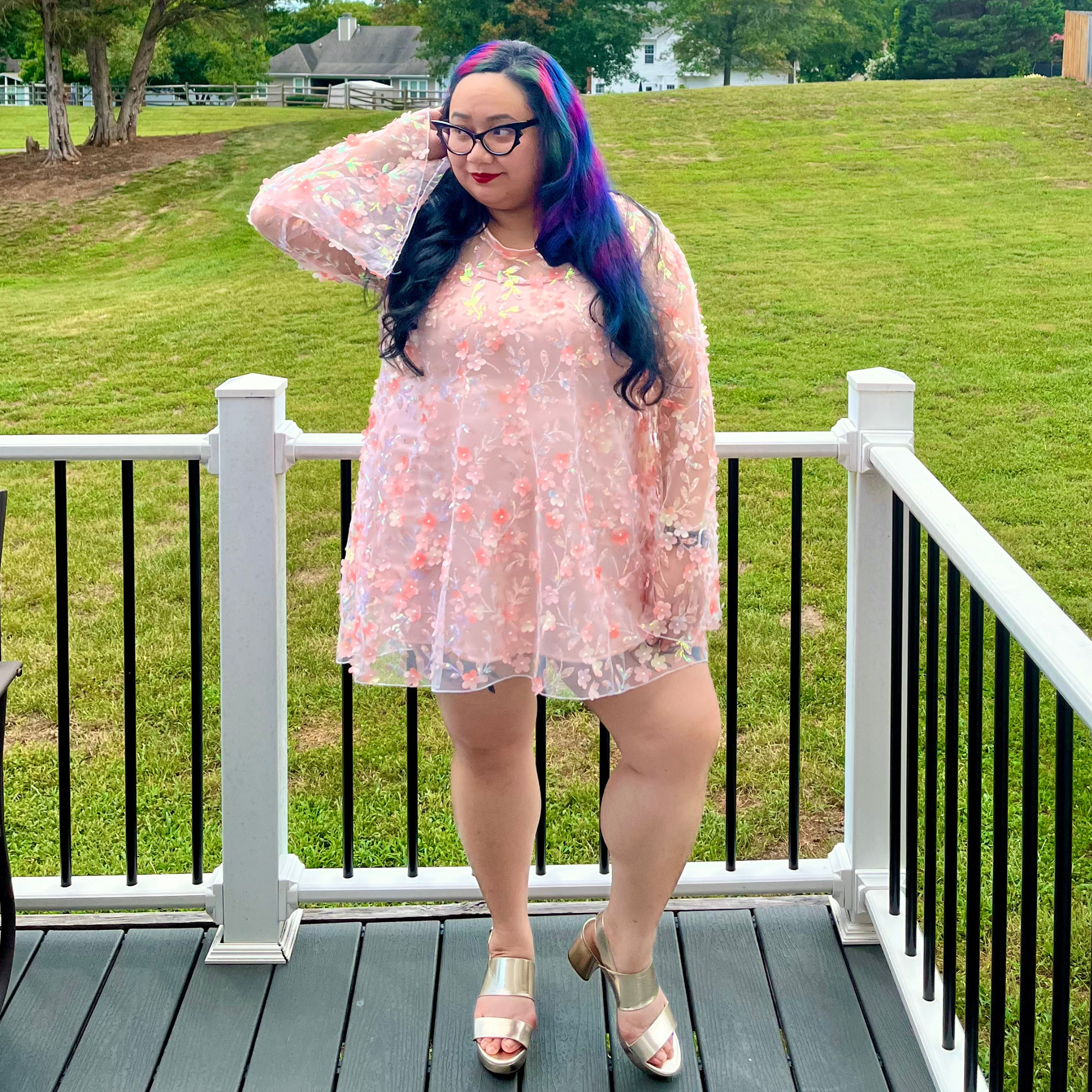 A woman stands on a wooden deck wearing a sheer light pink floral long-sleeve mini dress and metallic rose gold sandals, with a grassy lawn in the background.