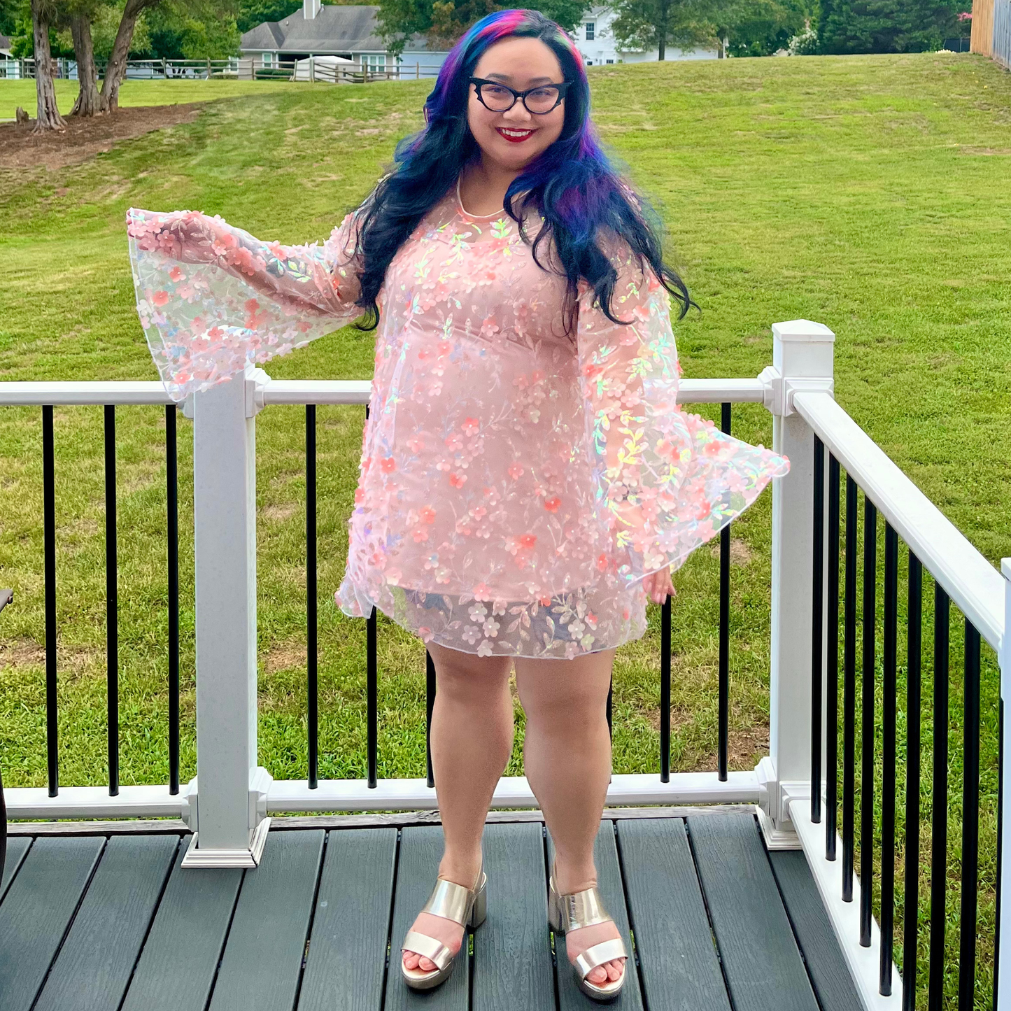 A woman with long blue and purple hair stands on a black deck railing wearing a sheer light pink floral long-sleeve tunic top with flared bell sleeves and a matching short skirt. The background shows a green grassy yard.