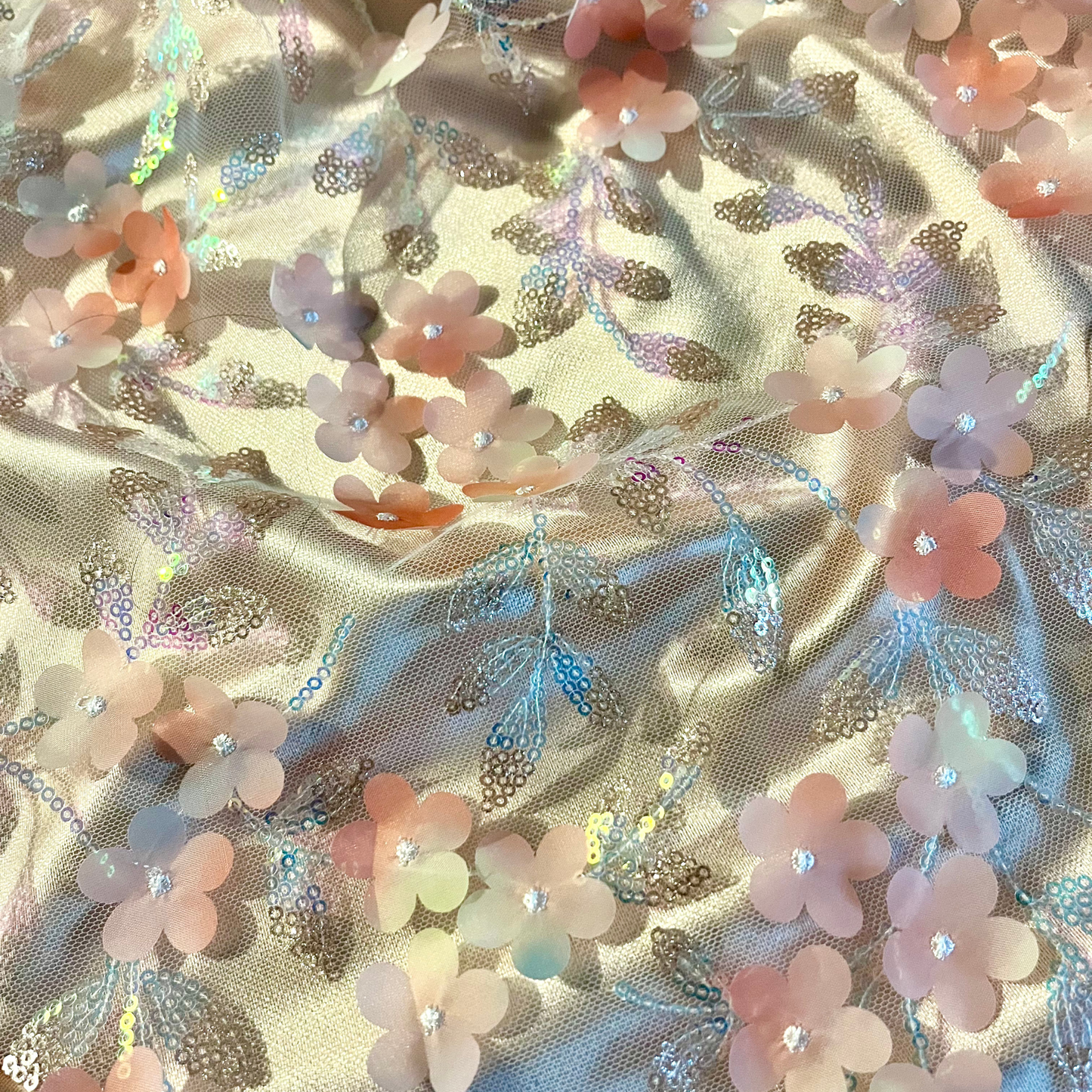 A close-up of iridescent floral fabric with peach, pink, and lavender flower appliqués, shimmering metallic leaves, and small rhinestone centers on a light beige satin-like background.