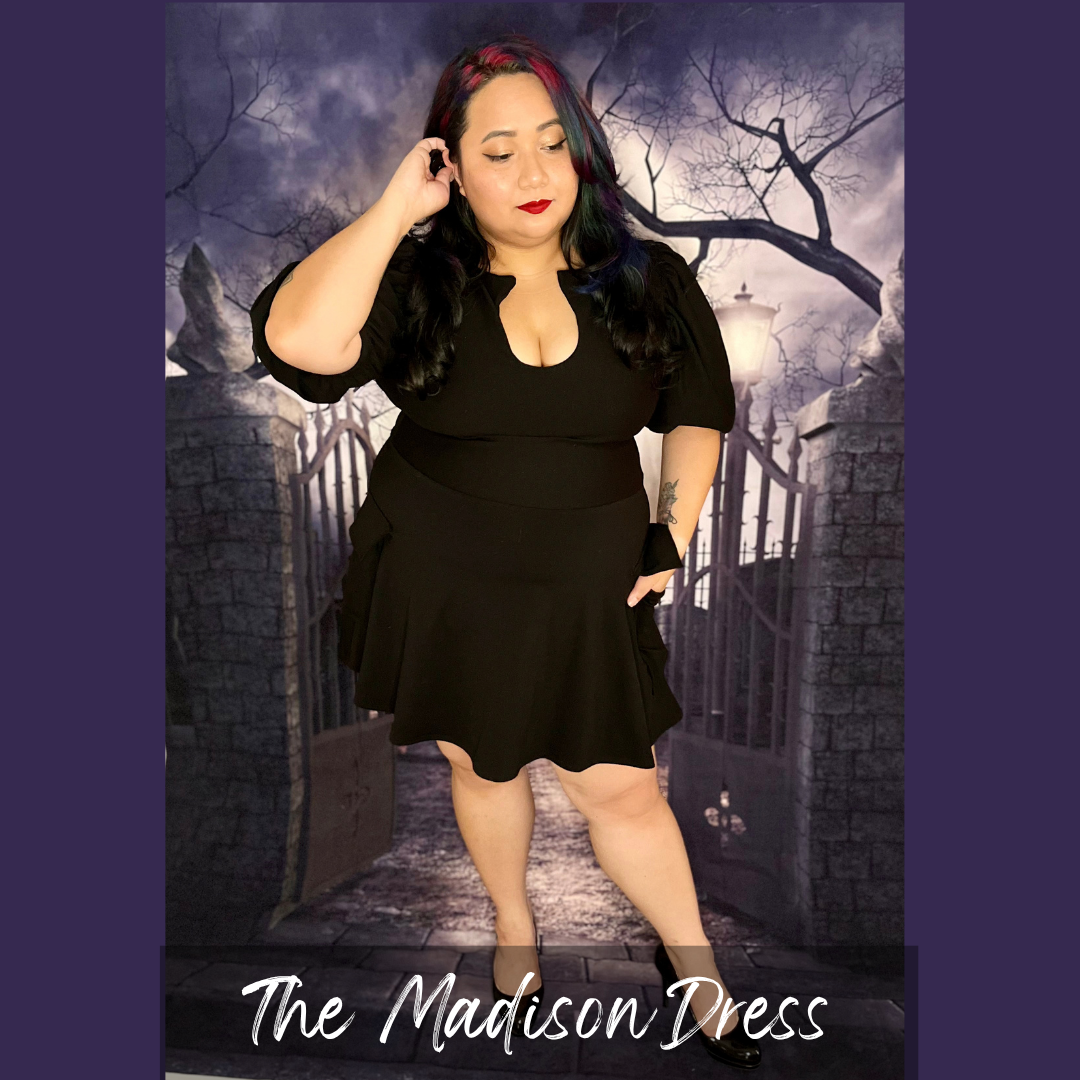 A woman modeling a black short-sleeve dress with a scoop neckline and flared skirt, standing in front of a gothic-themed backdrop with a stone gate and bare tree. The text 'The Madison Dress' appears at the bottom.