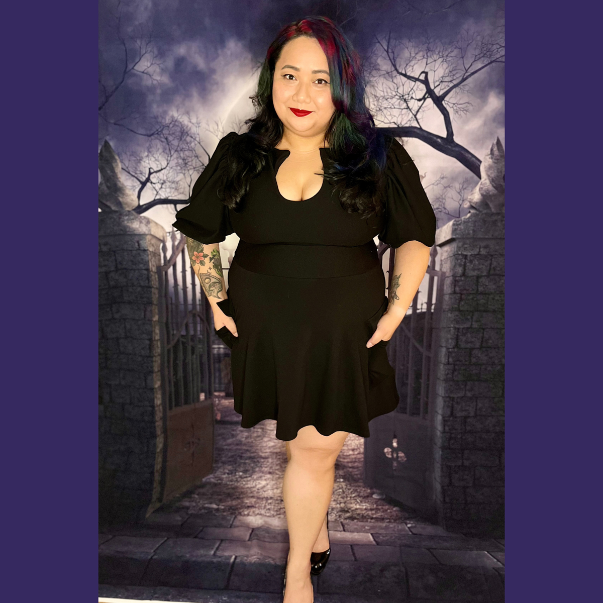 A woman modeling a black short-sleeve dress with puff sleeves and a flared skirt, standing in front of a gothic-themed backdrop of a stone gate and bare trees.