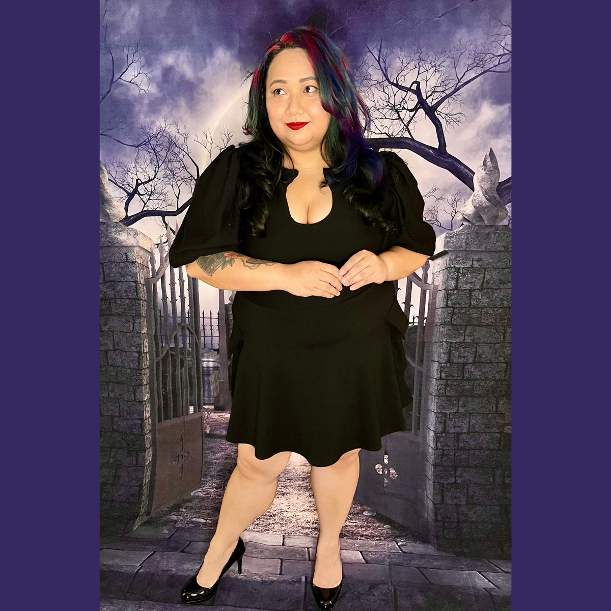 A woman modeling a black short puff-sleeve dress with a scoop neckline, paired with black high heels, standing in front of a gothic-themed backdrop of a stone gate, bare tree, and cloudy sky.