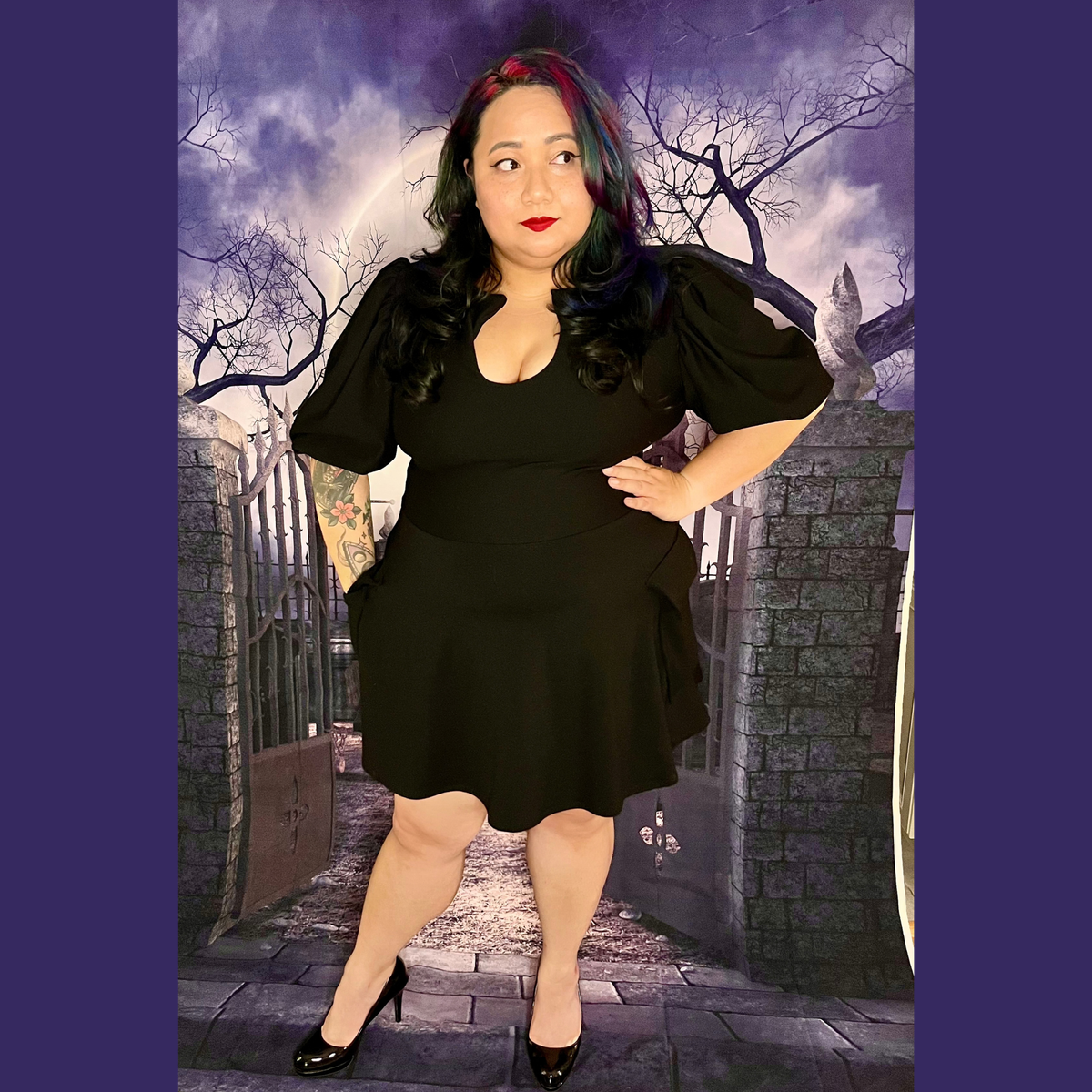 A woman modeling a black short-sleeve dress with puff sleeves and a flared skirt, standing in front of a gothic-themed backdrop featuring a stone gate, bare trees, and a cloudy sky.