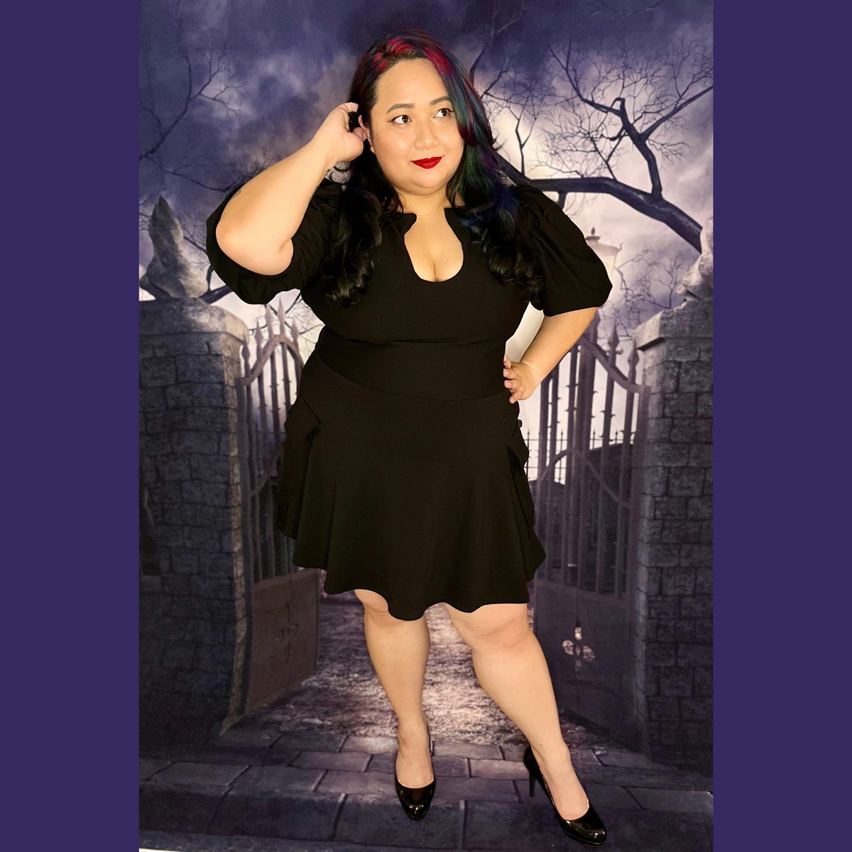 A woman modeling a black short-sleeve mini dress with puff sleeves and a scoop neckline, standing in front of a gothic-themed backdrop depicting a stone gate and bare trees.