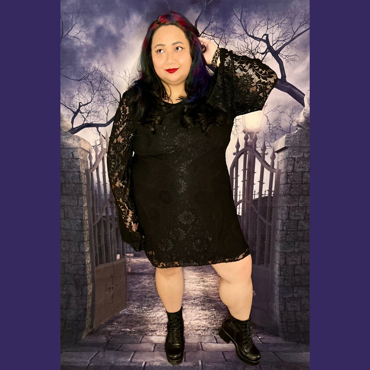 A woman modeling a black lace long-sleeve mini dress with black lace-up ankle boots, posed in front of a gothic-themed backdrop depicting a stone gate and a cloudy night sky.