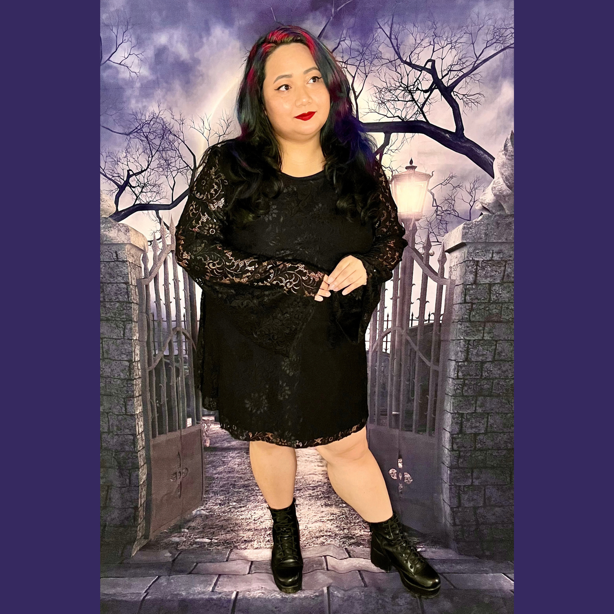 A woman modeling a black lace long-sleeve mini dress with black lace-up ankle boots, posed in front of a gothic-themed backdrop depicting a stone gate, brick walls, and a cloudy night sky.