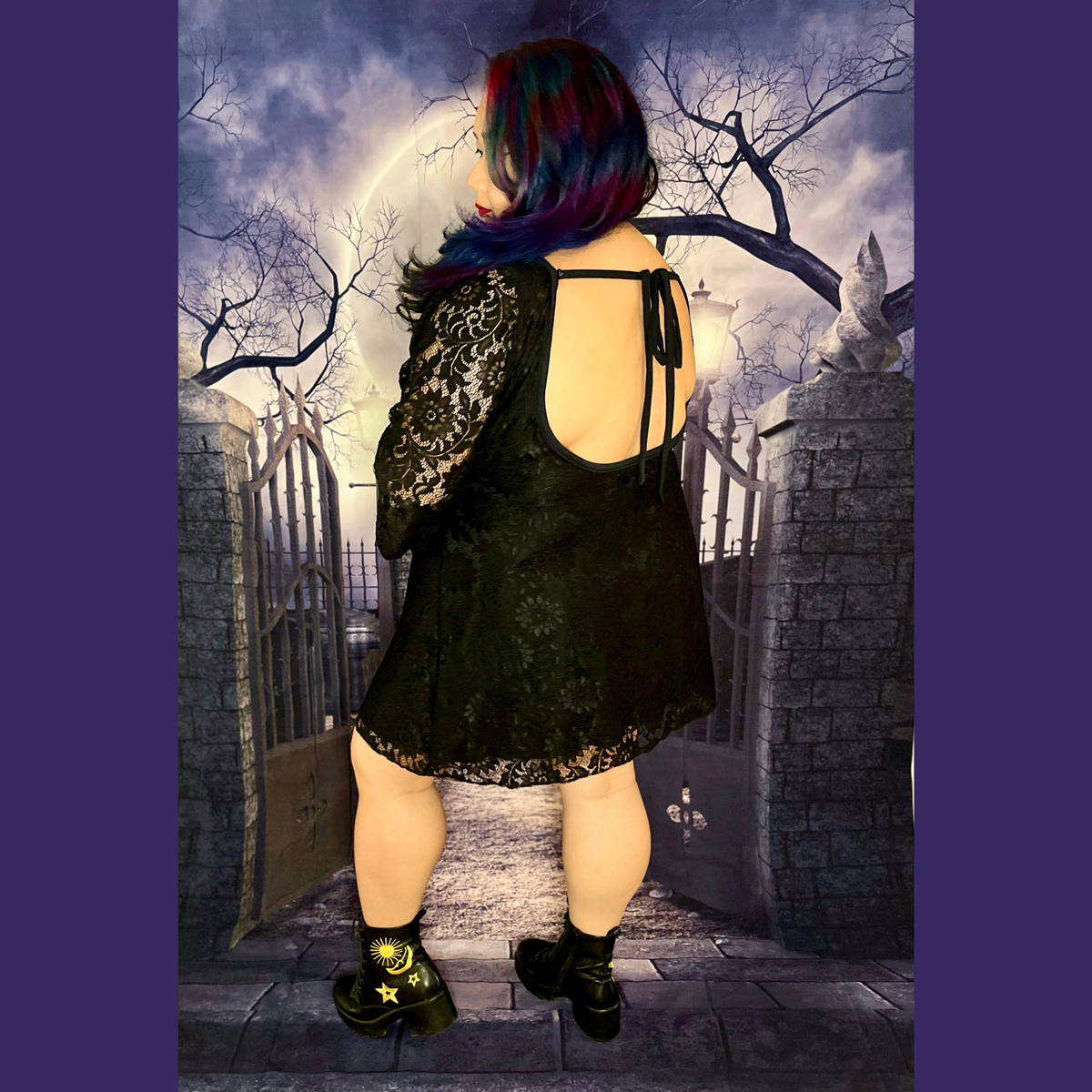 A person shown from behind wearing a black lace dress with long sheer sleeves and black ankle boots, standing in front of a gothic stone gate and full moon backdrop.