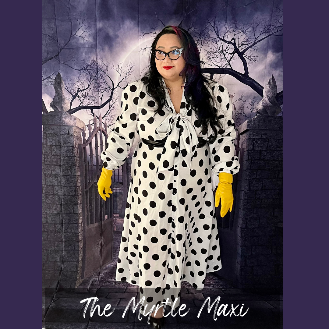 A person wearing a long-sleeve white polka-dot maxi dress with a tie neckline, styled with bright yellow gloves and glasses, standing in front of a spooky gothic gate backdrop. The text 'The Mirtle Maxi' is visible at the bottom.